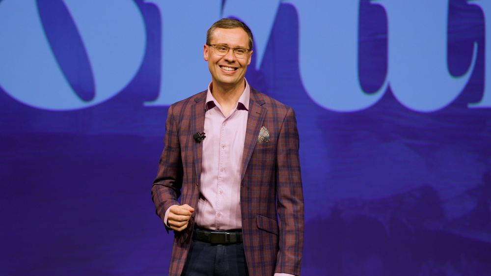Joshua Berry smiling on stage in a plaid jacket, in front of a bright blue screen