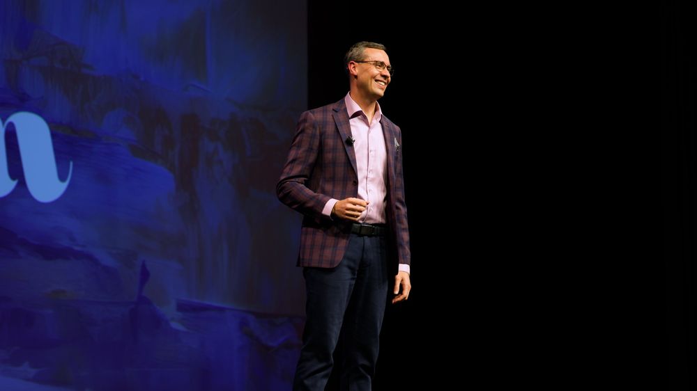 Wide shot of Joshua Berry on stage beside a large blue screen, with dark space to the right