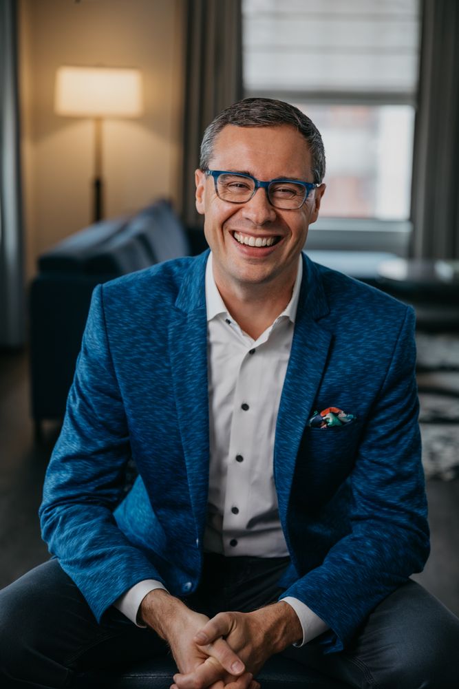 Joshua Berry laughing, seated in a blue jacket and blue glasses