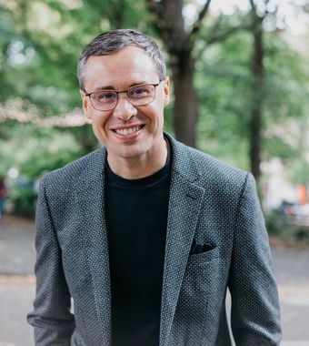 Joshua Berry smiling at the camera, in a grey blazer on a tree-lined street