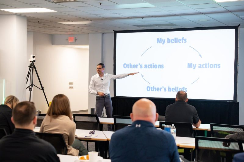 Joshua leading a workshop, pointing to a slide as a team listens from their tables